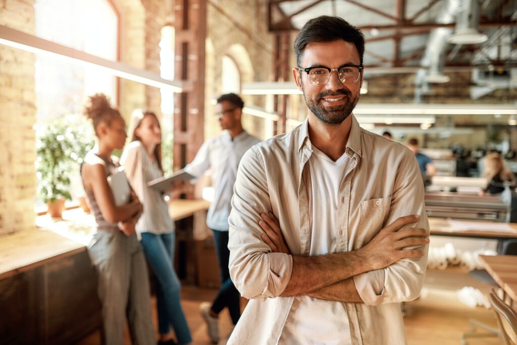 Great working day. Young bearded man in casual clothes keeping arms crossed and looking at camera